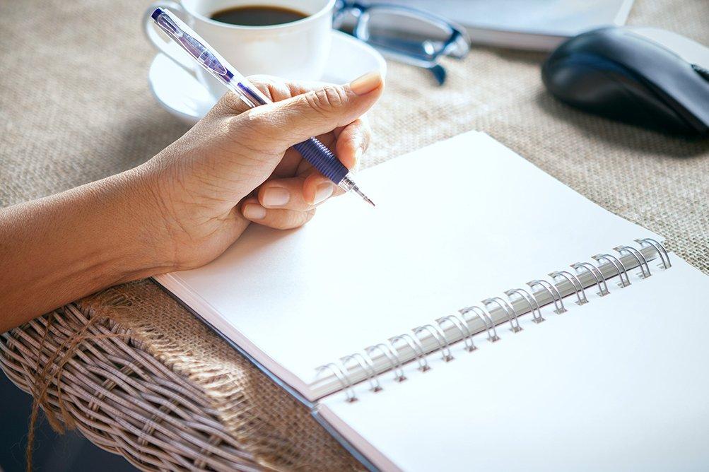 A person's left hand holding a pen over a blank page in their notebook