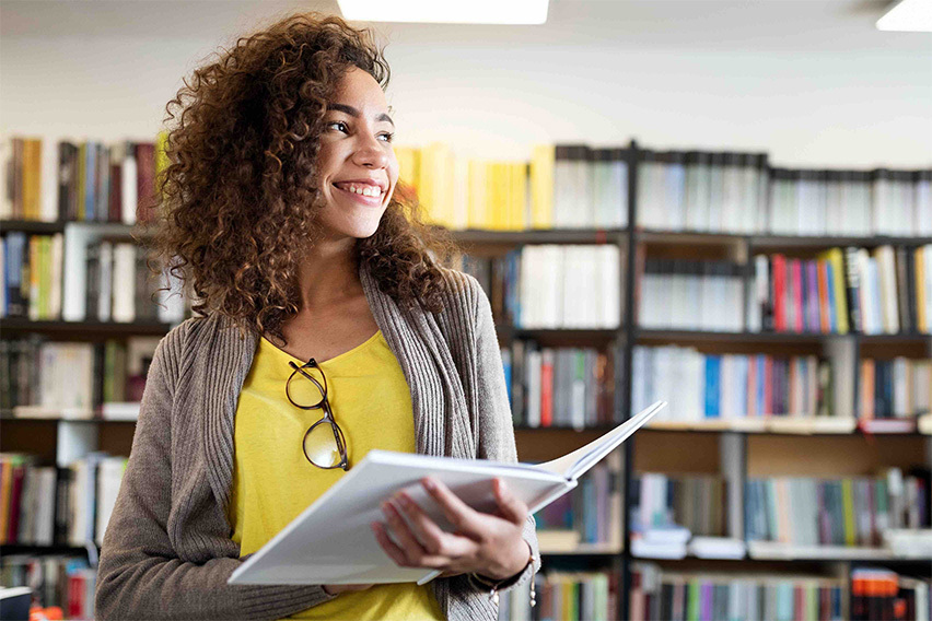 Girl in a yellow shirt with glasses on her shirt holds open a book. She is a UX design student about to graduate and is studying in a library.