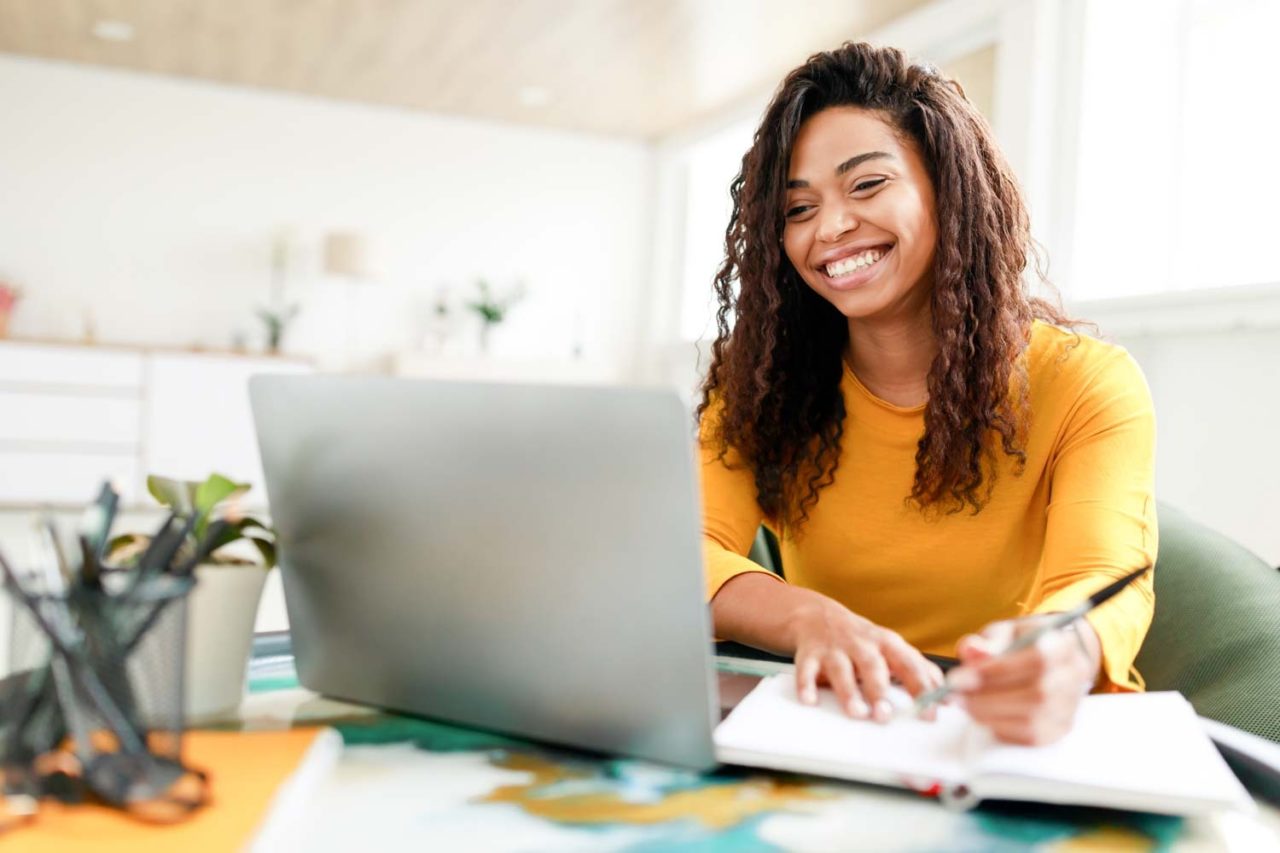 A young woman smiles as she sits at a desk with a laptop and notepad as she works on her UX portfolio.