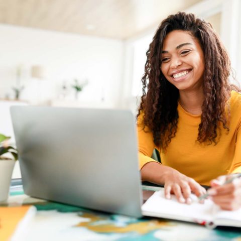 A young woman smiles as she sits at a desk with a laptop and notepad as she works on her UX portfolio.