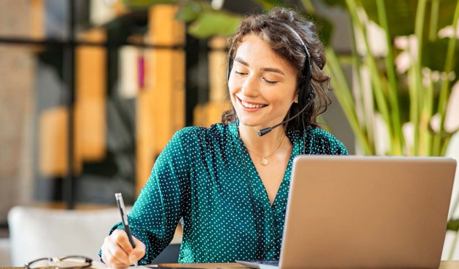 An image of a woman with a headset smiling and writing down notes as she talks to a customer in a call centre.