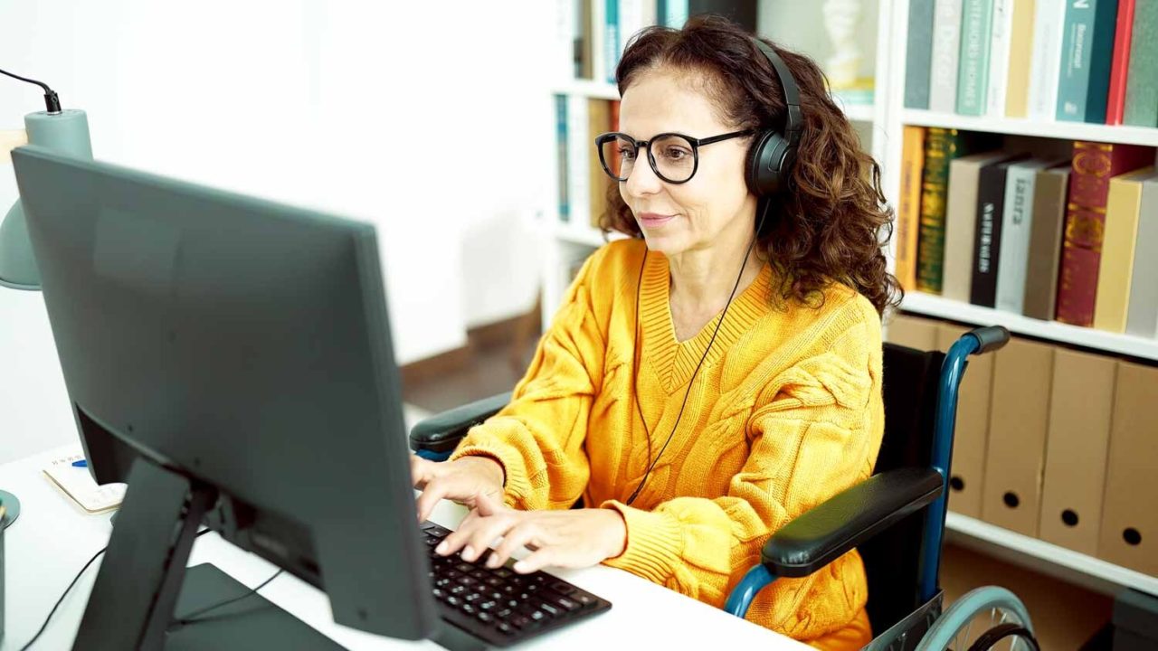 A woman with a yellow shirt is sitting at a computer desk using assistive technology to conduct a user testing session. She has curly hair and headphones on. 
