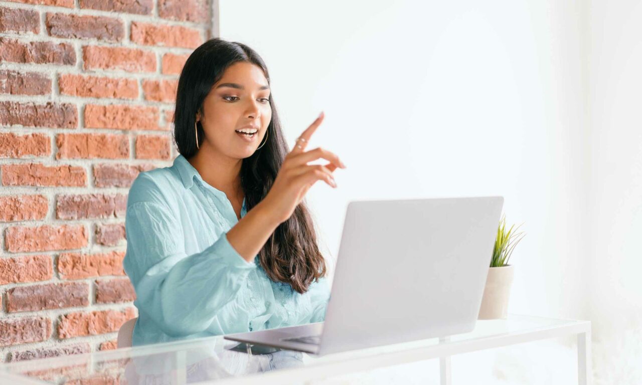 Image of a woman working remotely, sitting at a desk with her laptop, gesturing and speaking during a video call