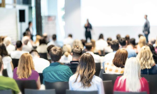 A photo of a conference room with audience members sitting in chairs watching a presentation.