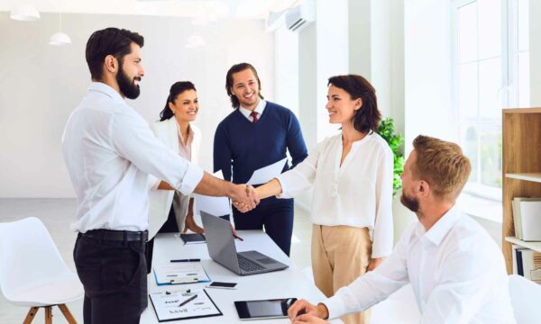 A group of UX Professionals standing around a table in a business environment, shaking hands.