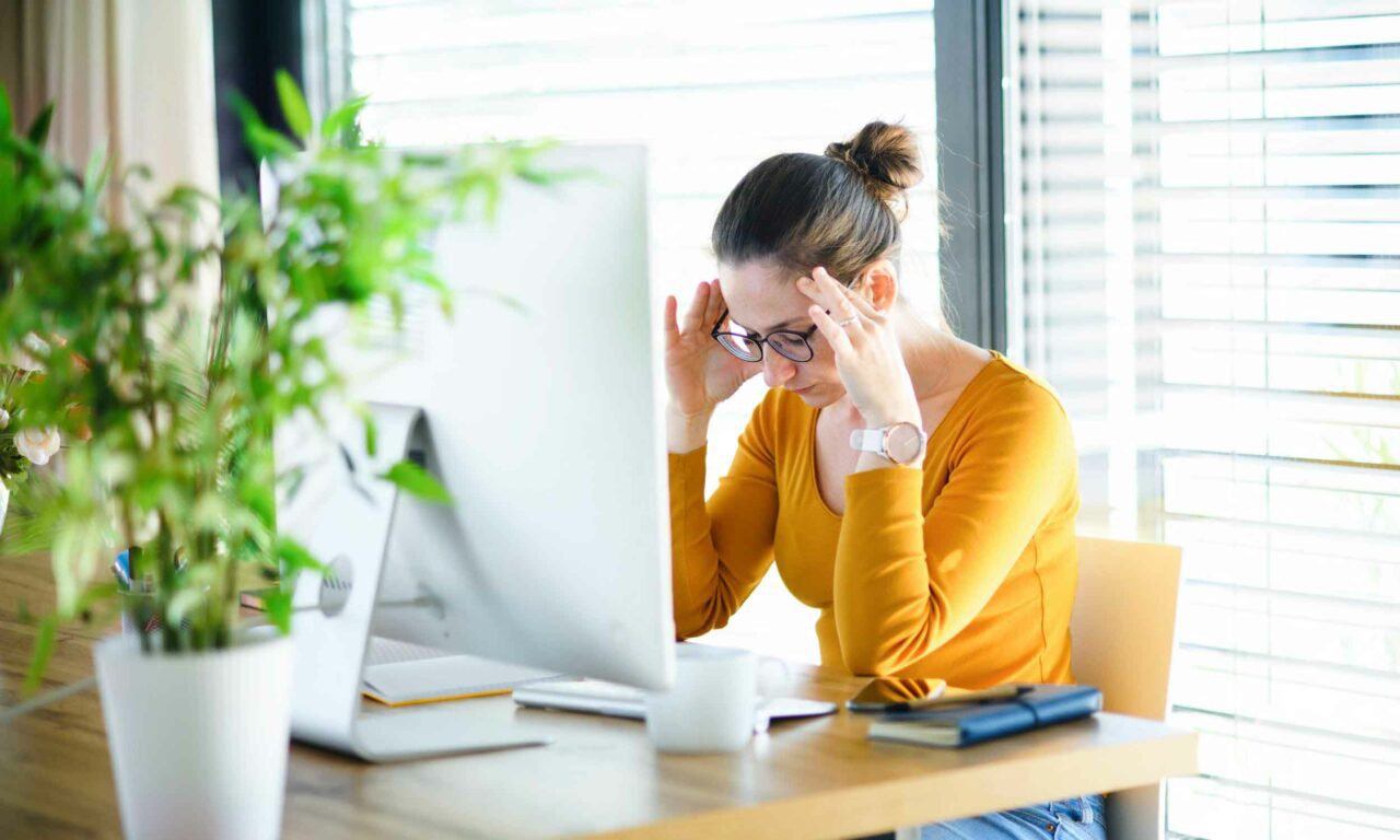 A UX designer sits at her desk, head in her hands, looking overwhelmed. She is wearing a yellow long sleeved top and glasses. 