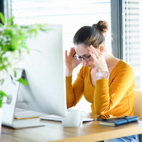 A UX designer sits at her desk, head in her hands, looking overwhelmed. She is wearing a yellow long sleeved top and glasses.