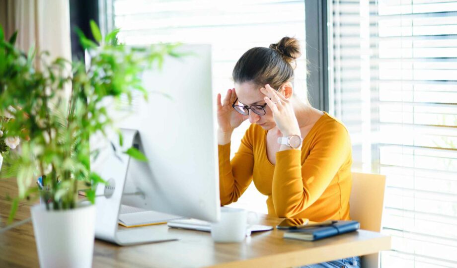 A UX designer sits at her desk, head in her hands, looking overwhelmed. She is wearing a yellow long sleeved top and glasses.