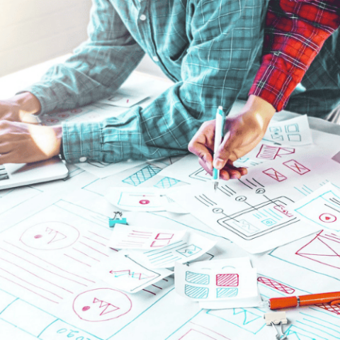 A pair of designers working together on a project. The left most person has a blue shirt and is using a leptop. The person on the right is wearing red and has some design wireframes scattered in front of them.