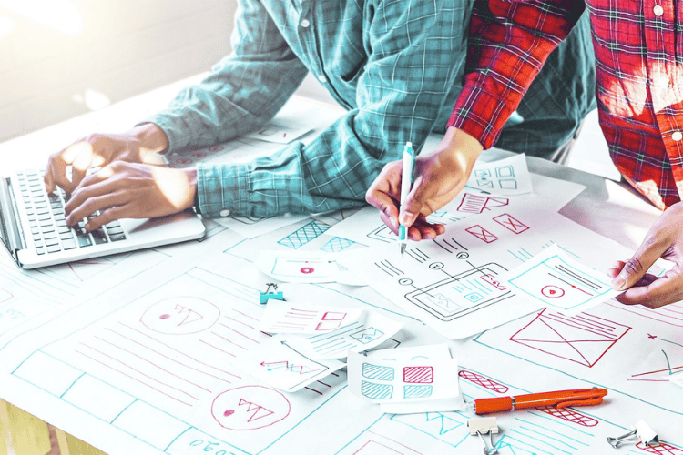 A pair of designers working together on a project. The left most person has a blue shirt and is using a leptop. The person on the right is wearing red and has some design wireframes scattered in front of them.