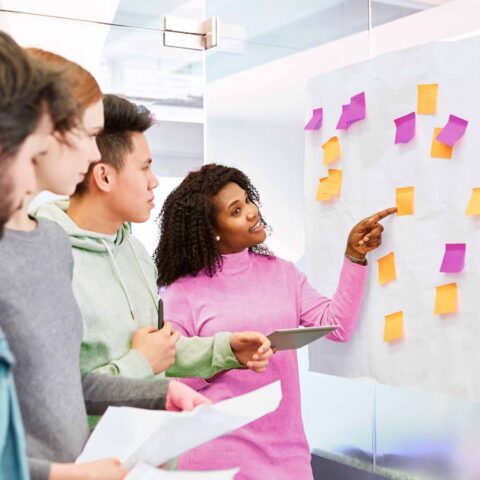 Designers and researchers running a workshop "How Might We" statements and pointing at sticky nots on a whiteboard.