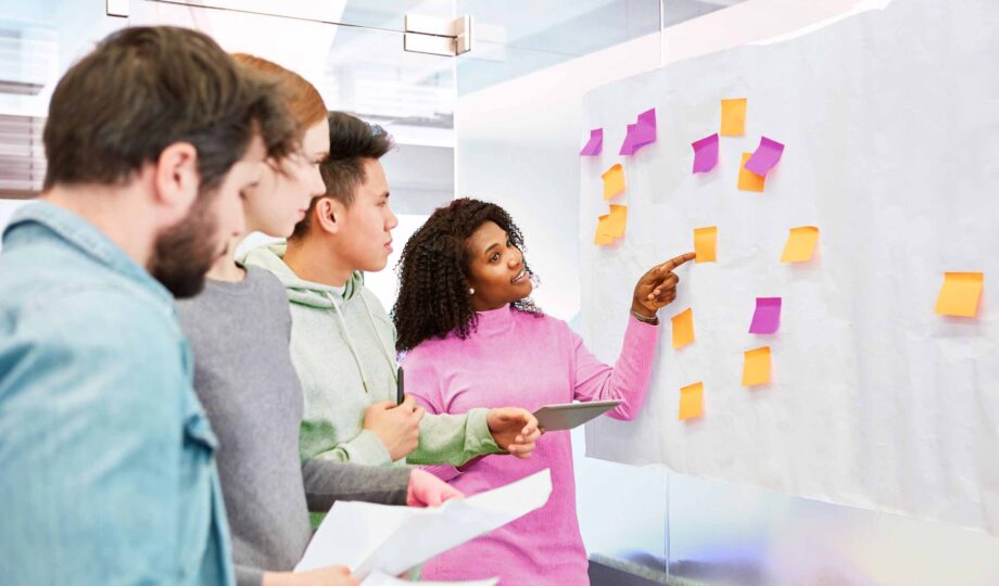 Designers and researchers running a workshop "How Might We" statements and pointing at sticky nots on a whiteboard.