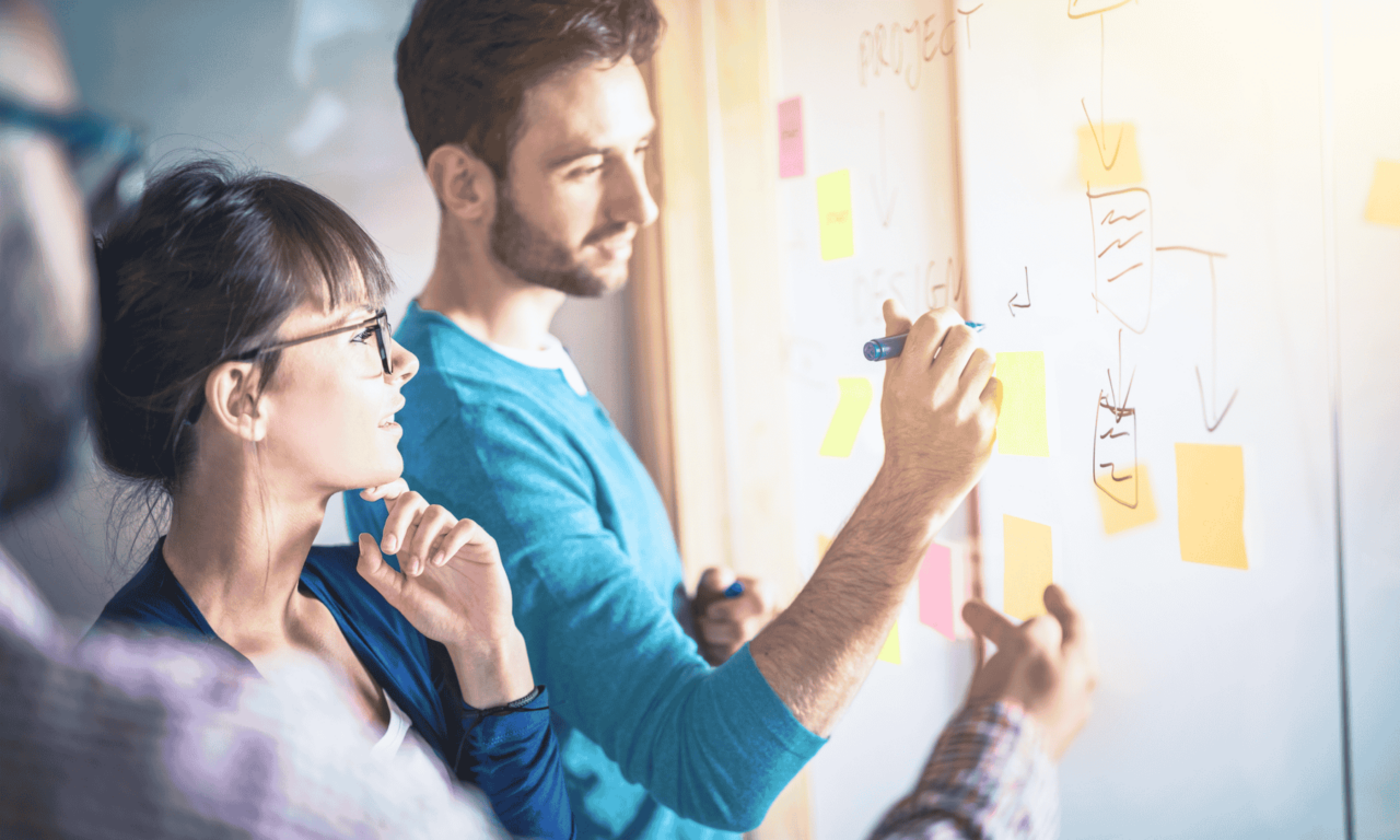 A UX designer and a UX researcher brainstorming at a whiteboard as they practice a human-centered design (HCD) approach.