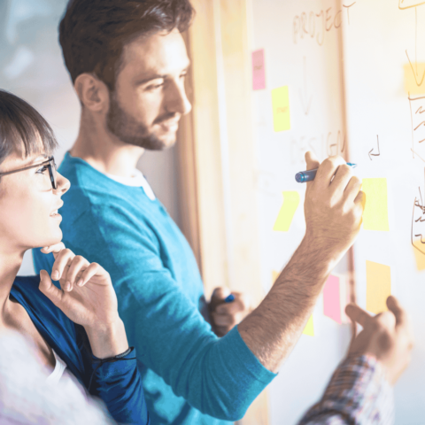 A UX designer and a UX researcher brainstorming at a whiteboard as they practice a human-centered design (HCD) approach.