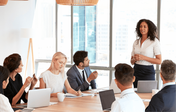 A woman standing and presenting to a group of coworkers seated around a conference table in a bright modern office