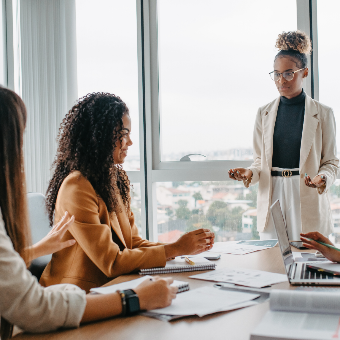 A professional office meeting: three women are seated around a conference table with laptops, notebooks, and documents.