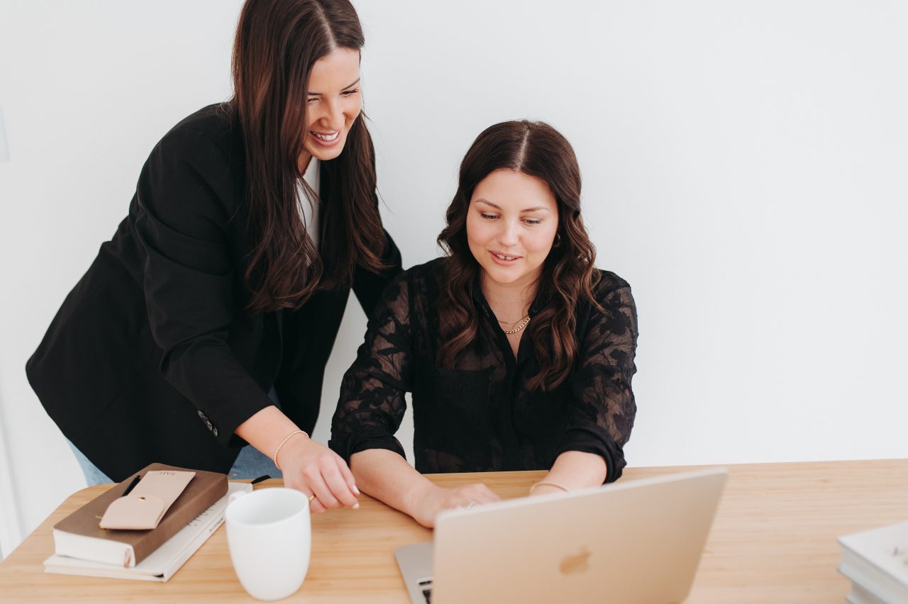 Two women working together at a desk, one sitting at a laptop while the other stands beside her and points at the screen.