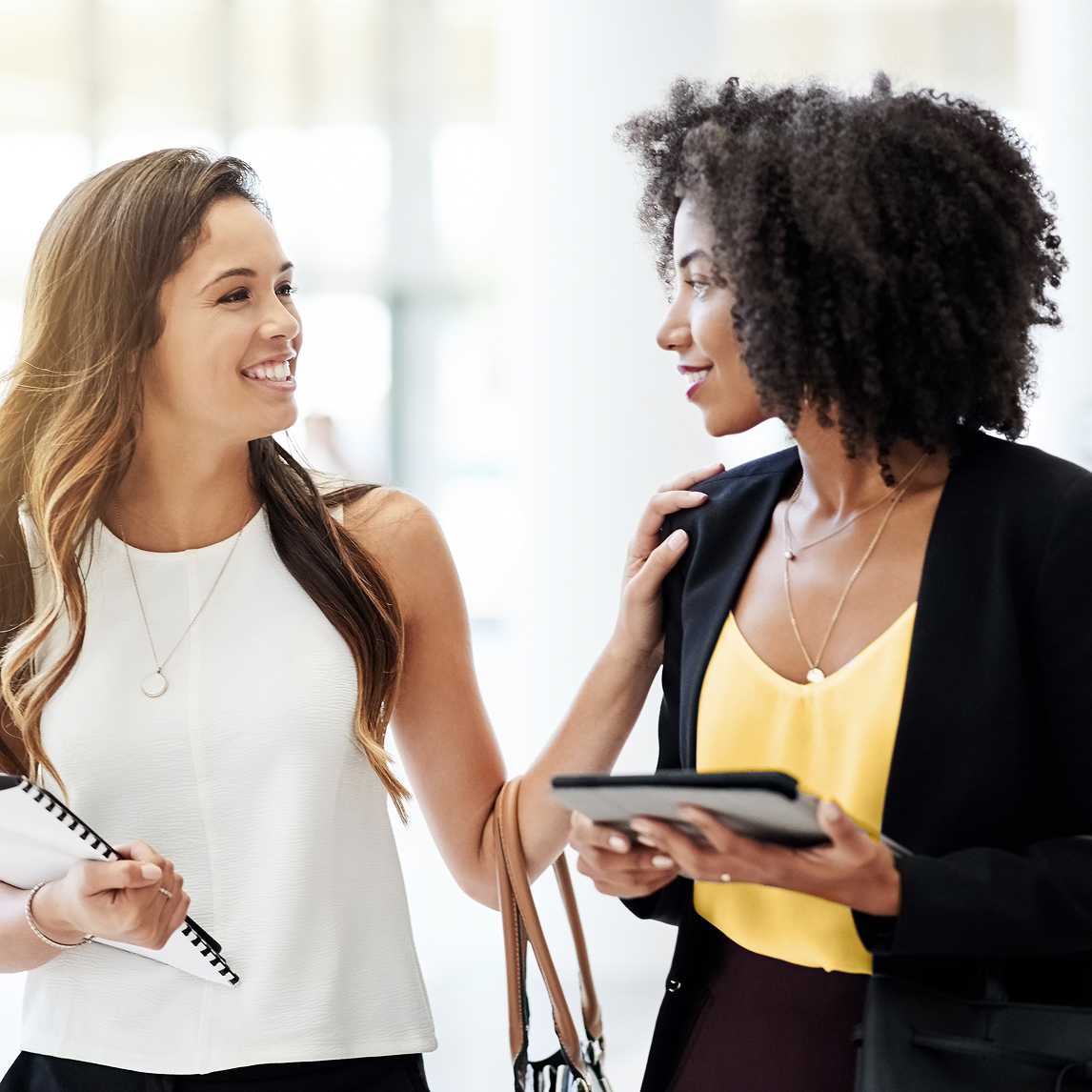 Two professional women walking and talking in a bright office hallway