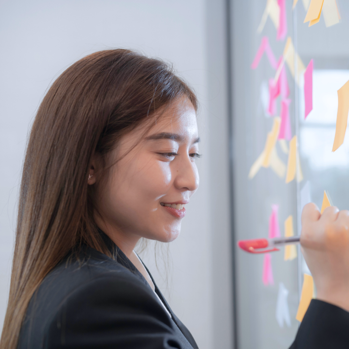 A young woman with long, straight brown hair, wearing a dark blazer over a white collared shirt, is standing in profile in front of a glass wall or whiteboard covered with colorful sticky notes (pink, yellow, and orange).