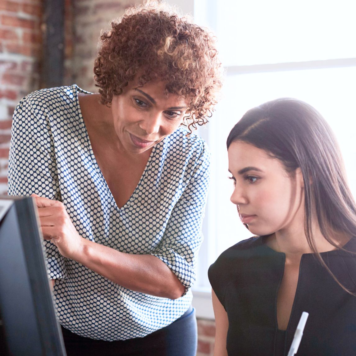 A professional office setting: a woman with short, curly reddish-brown hair wearing a blue-and-white polka-dot blouse is standing and leaning forward, pointing at or explaining something on a computer screen.