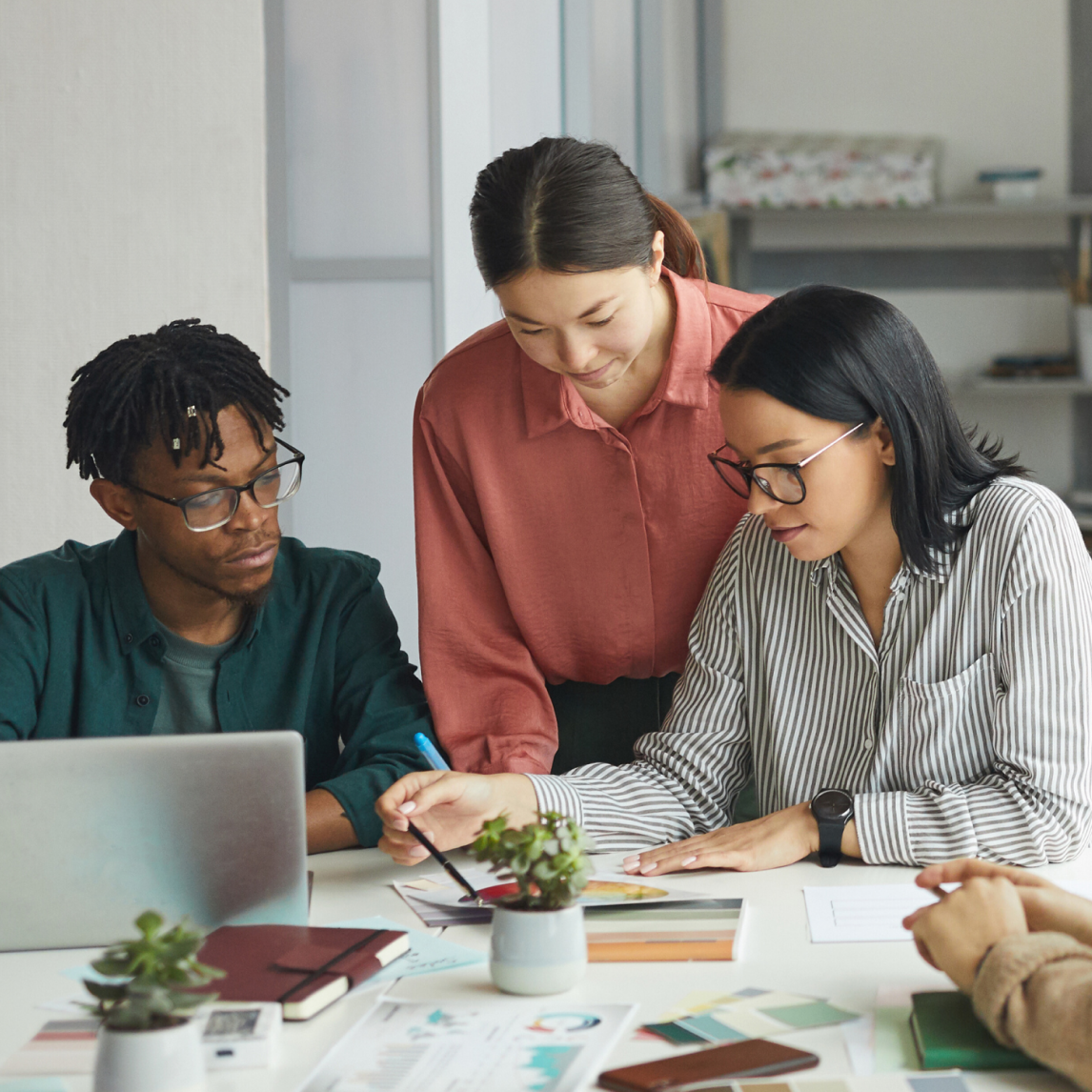Three colleagues collaborating around a table in a modern office. A woman in a coral blouse stands and leans over the table, pointing with a pen at documents. A man with short dreadlocks, wearing glasses and a green shirt, sits on the left looking at a laptop, while a woman with straight black hair and glasses, dressed in a striped shirt, sits on the right reviewing papers. The table is covered with notebooks, printed charts, color swatches, small potted plants, and scattered documents, creating a focused and creative team-working atmosphere.