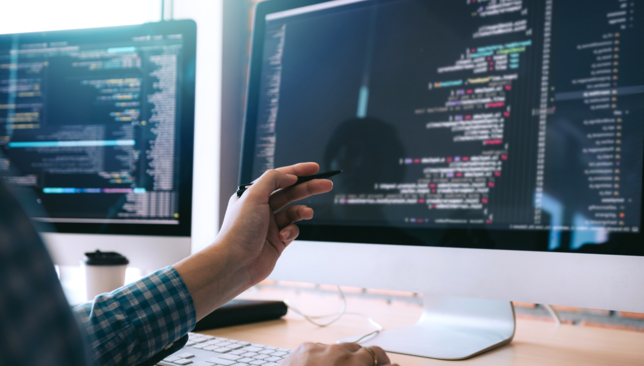 Software developer for a networking IT system, pointing at code on dual monitors in a modern office workspace