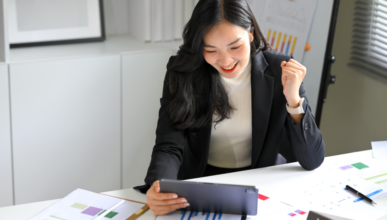 Excited businesswoman checking sales analytics on tablet at office desk with charts, celebrating growth results