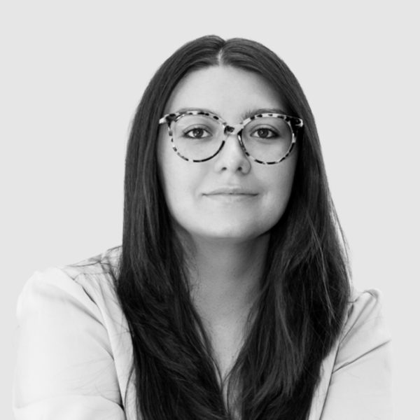 Professional headshot of woman with long dark hair wearing tortoiseshell round glasses on a light gray studio background