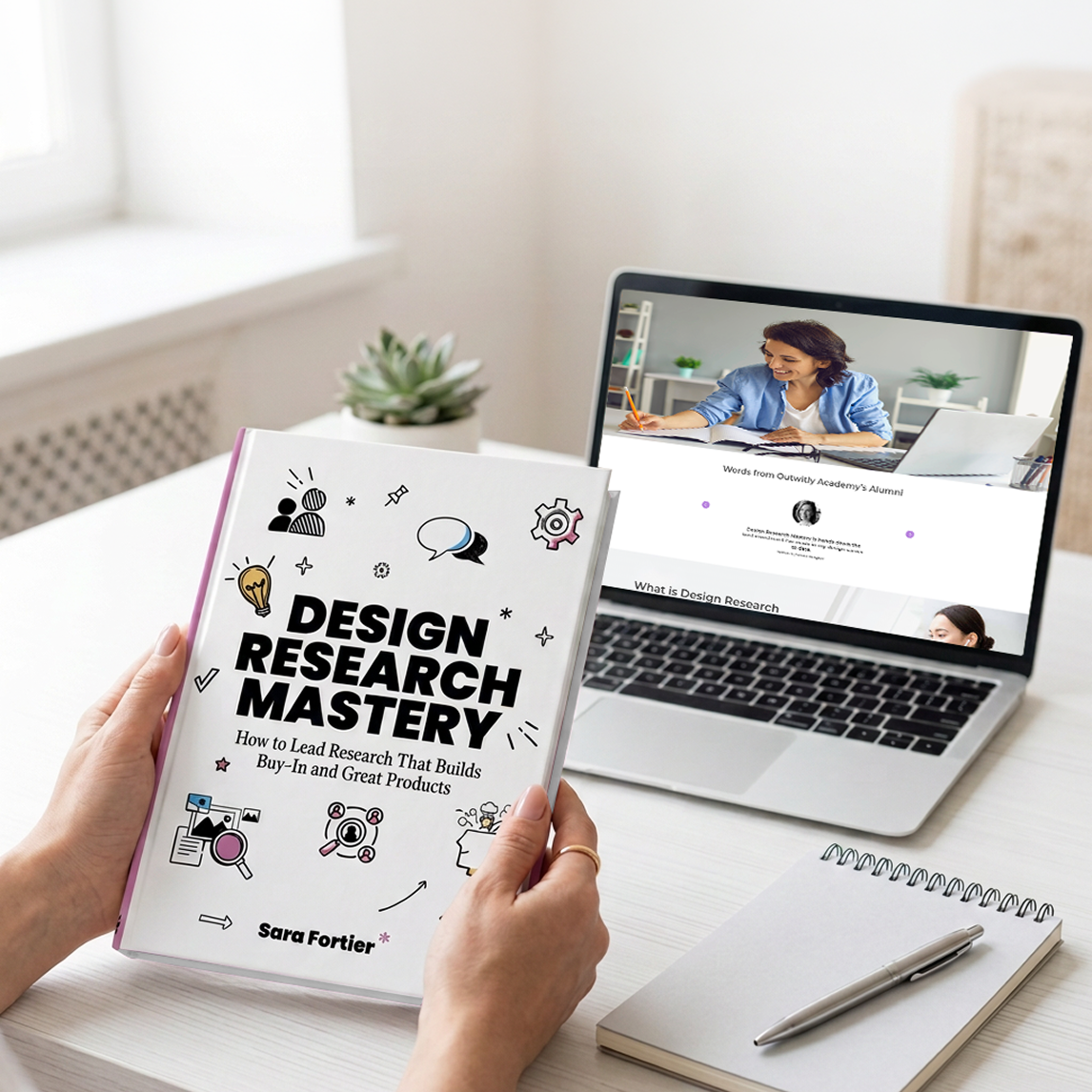 Hands holding “Design Research Mastery” book beside laptop and notebook on desk, promoting UX research training.